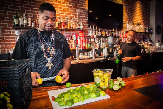 (L-R) A bar-back Milan Hawthorne “BeatsMe” cuts a full basket of limes preparing for drinks while his colleague, the bartender Stephen Sloper makes a drink at the Liege Spirits Lounge in Oakland on Friday, July 24, 2015. (Photo by Ekevara Kitpowsong/Bay News Rising)