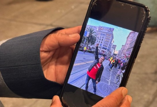 Two hands hold a smartphone, which displays an image of a man holding up his fist on a city street